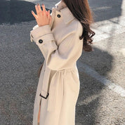 Cappotto beige elegante da donna, lungo con cintura, bottoni neri, indossato all'aperto. Moda autunnale, stile casual chic.
