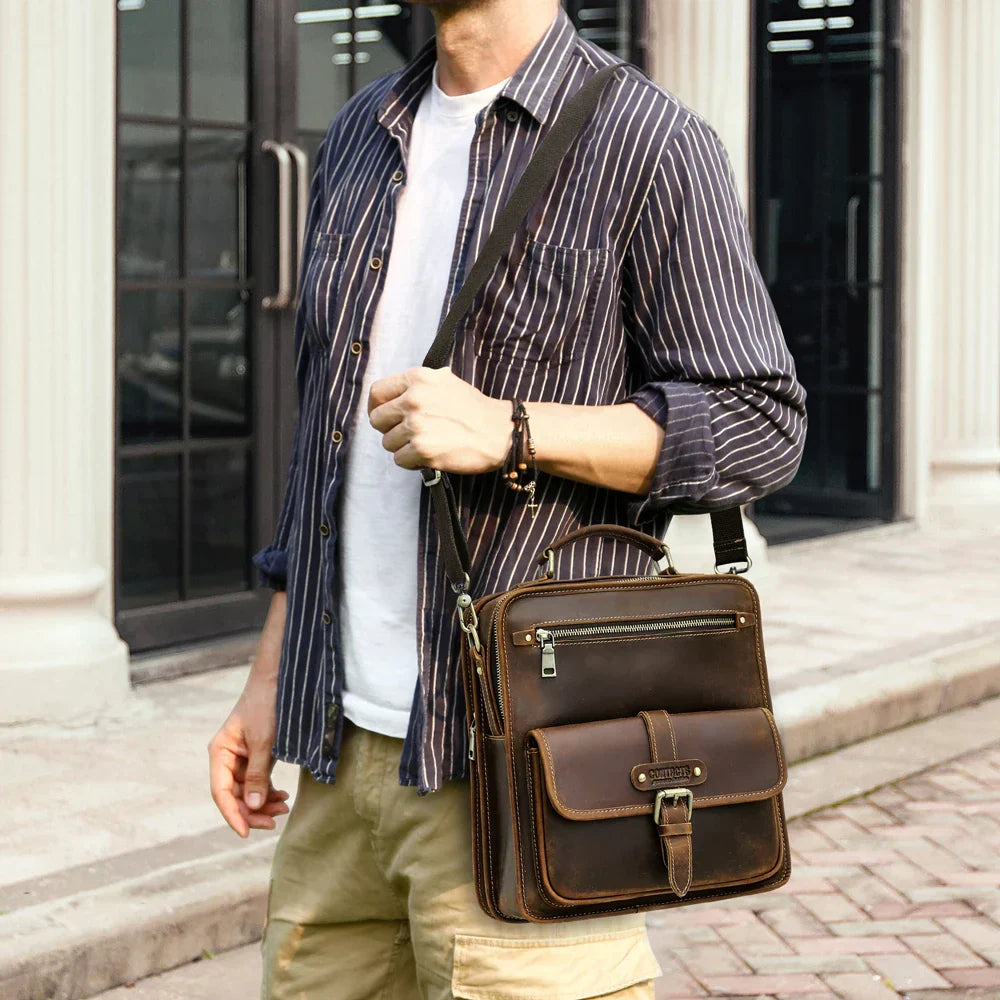 Uomo con borsa a tracolla in pelle marrone, tasca frontale con zip, abbigliamento casual. Accessorio moda maschile elegante e pratico.
