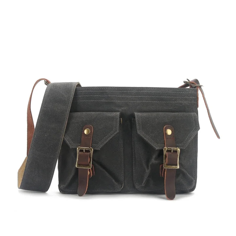 Borsa a tracolla in tela cerata nera con doppie tasche frontali, fibbie in pelle marrone e tracolla regolabile. Stile vintage, resistente e versatile.