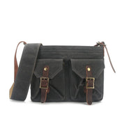 Borsa a tracolla in tela cerata nera con doppie tasche frontali, fibbie in pelle marrone e tracolla regolabile. Stile vintage, resistente e versatile.