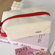 Borsa da toilette in tessuto beige con cerniera rossa, posizionata su un libro rosa. Design moderno, ideale per viaggi e organizzazione.