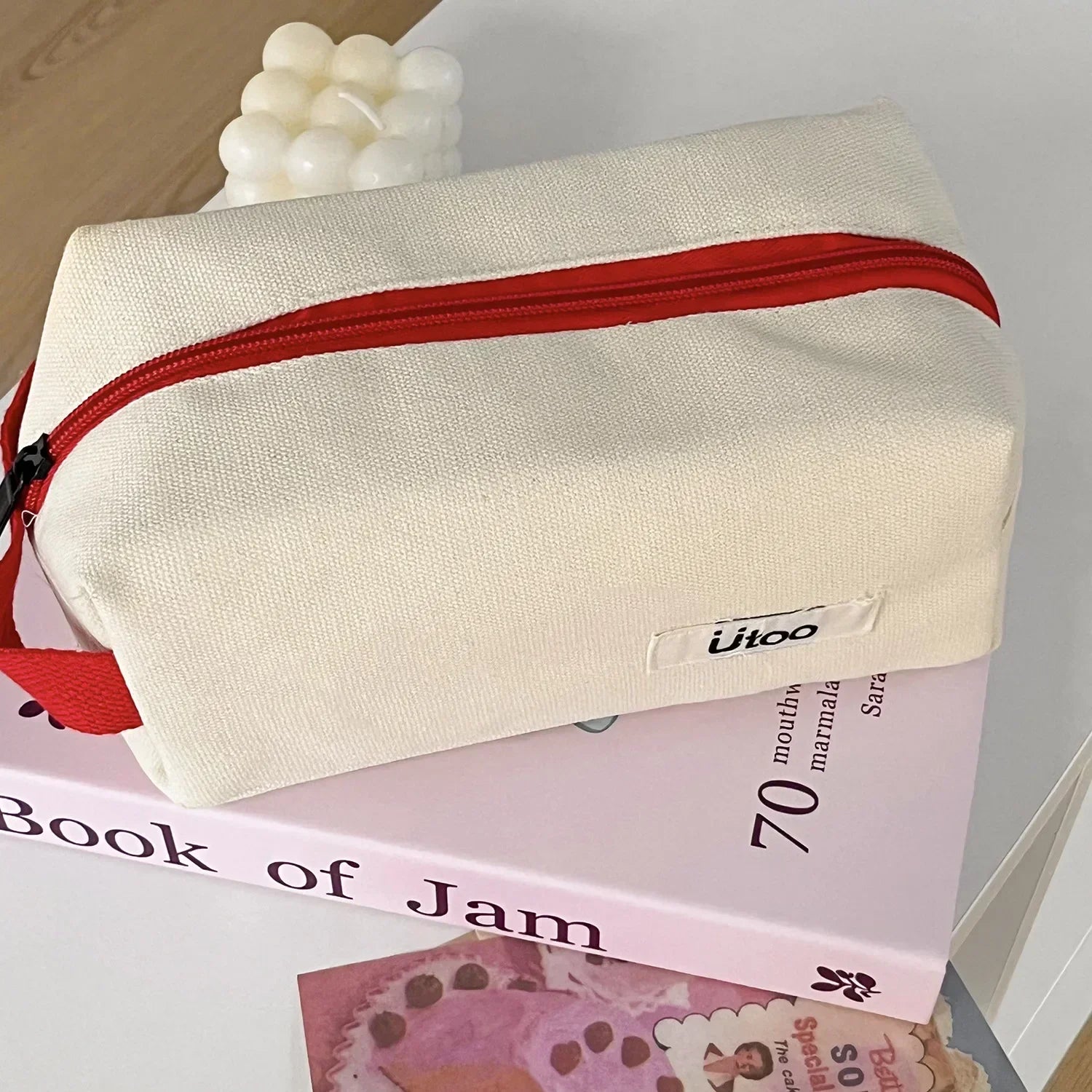 Borsa da toilette in tessuto beige con cerniera rossa, posata su un libro rosa. Design moderno, ideale per viaggi e organizzazione accessori.