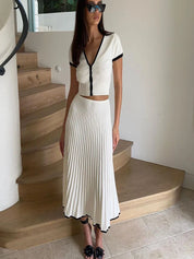 Donna con abito bianco a pieghe, dettagli neri, occhiali da sole, posa su scala interna. Moda elegante, abbigliamento estivo, stile chic.