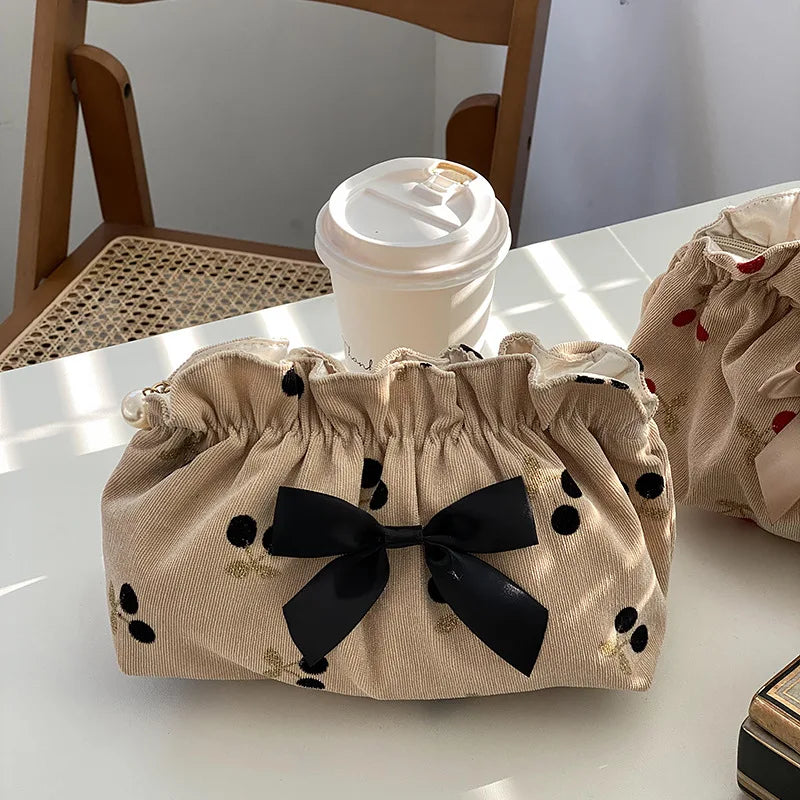 Borsa beige con fiocco nero e pois neri, accanto a un bicchiere di caffè su tavolo in legno. Accessorio moda elegante e pratico.
