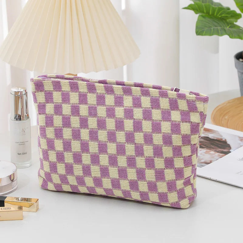 Borsa a scacchi viola e beige su tavolo bianco, accanto a lampada e piante. Accessorio moda elegante, ideale per organizzare cosmetici.