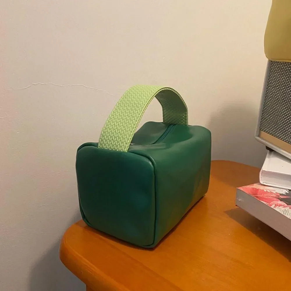 Borsa verde in pelle con manico intrecciato verde chiaro su tavolo in legno. Design moderno, accessorio moda elegante, ideale per ogni occasione.