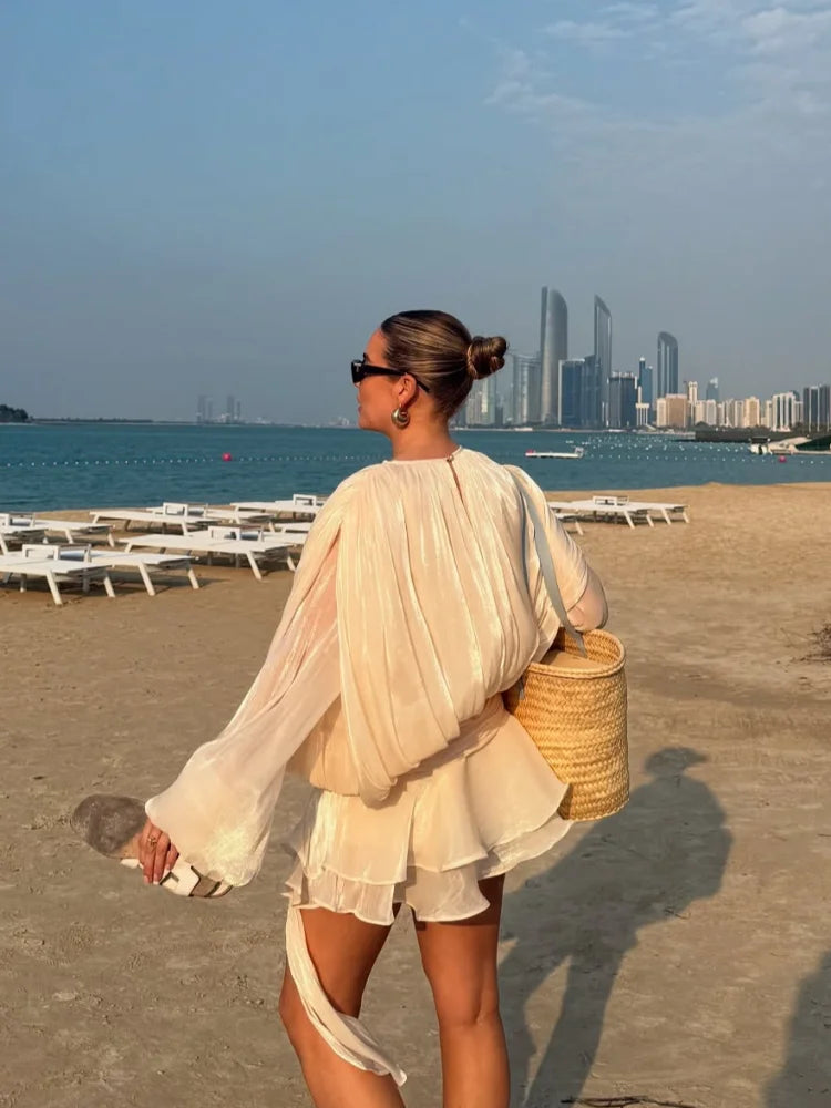 Donna elegante in abito estivo bianco su spiaggia, con borsa di paglia e occhiali da sole. Grattacieli sullo sfondo. Moda mare, look estivo chic.