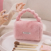 Borsa rosa in pelliccia sintetica con manico intrecciato, circondata da pennelli da trucco su un tavolo. Design elegante e moderno.