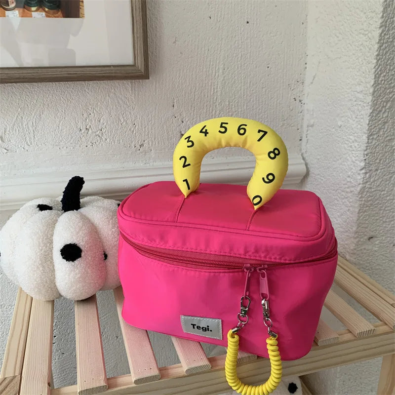 Borsa rosa con manico giallo numerato, design moderno, su scaffale in legno accanto a decorazioni bianche. Accessorio moda unico e colorato.