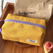 Astuccio giallo in tessuto con cerniera viola su tavolo di legno, marchio "ütoo". Sfondo con busta di carta marrone e scritta "happiness".