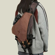 Zaino marrone a tracolla con borraccia nera, indossato da una persona con maglione grigio. Accessorio moda pratico per escursioni e viaggi.