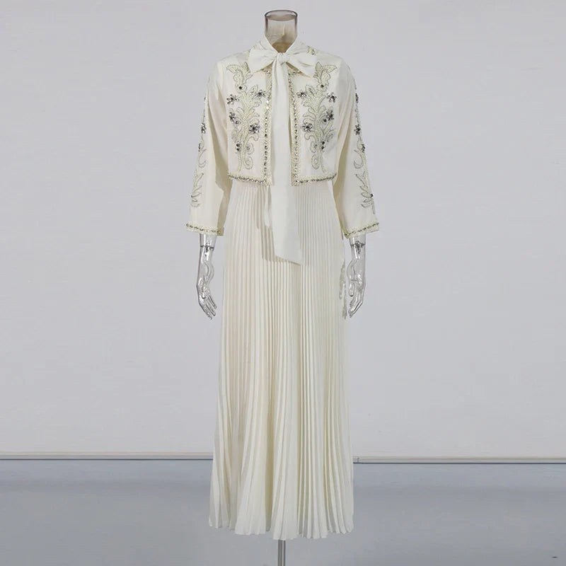 Abito lungo bianco elegante con ricami floreali, maniche lunghe e colletto a fiocco. Design plissettato, ideale per cerimonie e occasioni speciali.