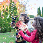 Bambini giocano con pistola sparabolle all'aperto, bolle di sapone colorate, divertimento in giardino, attività per bambini, giornata soleggiata.