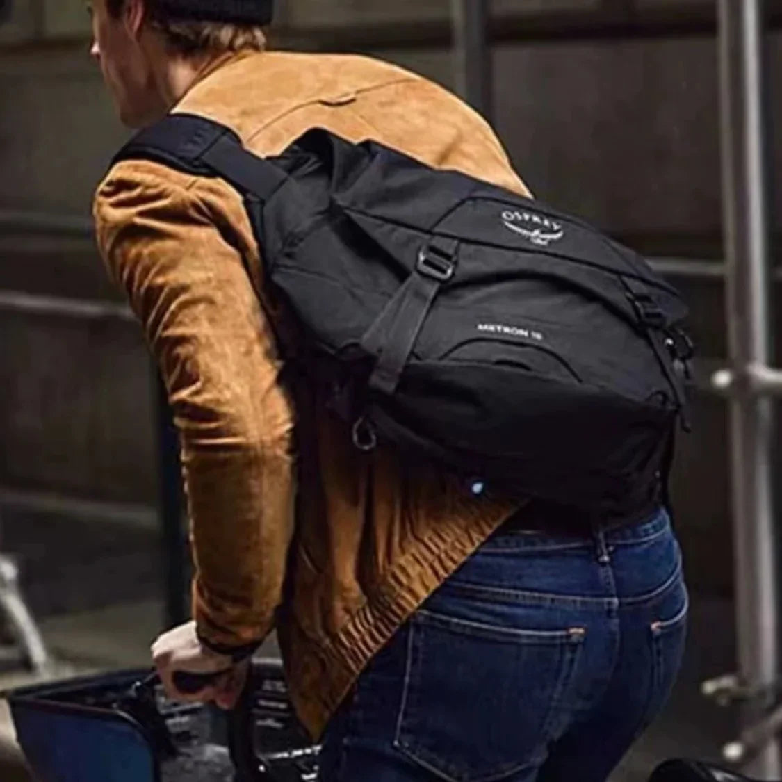Uomo con zaino nero Osprey Metron 18, giacca marrone e jeans, in bicicletta. Accessorio urbano, moda casual, pratico per pendolari.