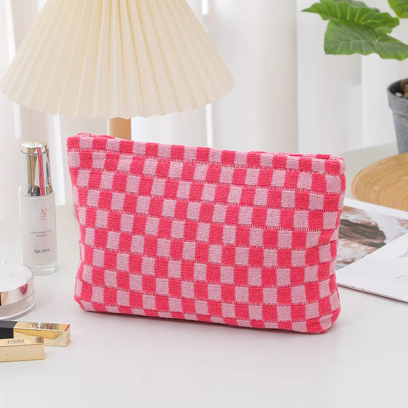 Astuccio rosa a quadretti bianchi su tavolo bianco, accanto a lampada e pianta. Design moderno, ideale per cosmetici o accessori.