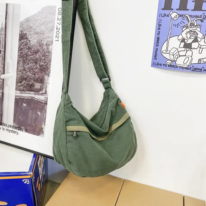 Borsa a tracolla verde in tessuto, appesa a un gancio su parete bianca. Design casual, ideale per uso quotidiano. Accessorio moda pratico e versatile.