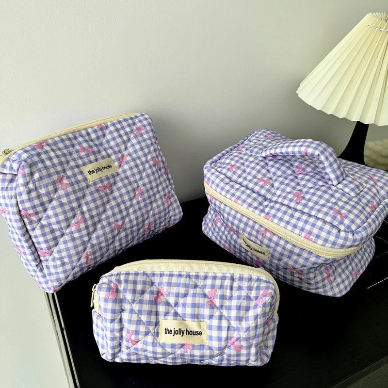 Set di tre beauty case a quadretti lilla e bianchi con dettagli rosa, marchio "the jolly house", su tavolo nero accanto a lampada bianca.