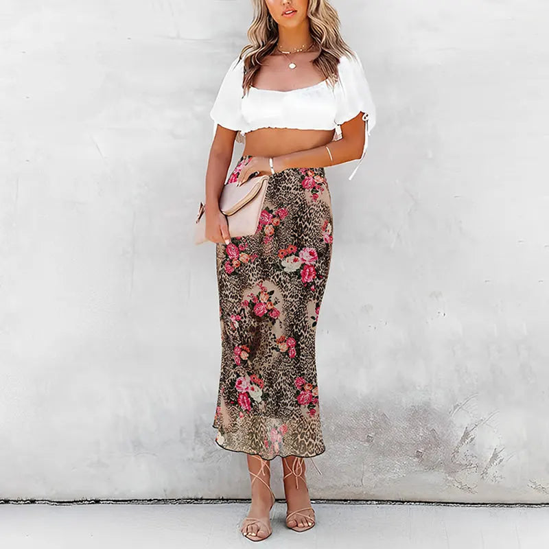 Donna con top bianco e gonna lunga floreale, sandali beige, borsa a mano rosa. Moda estiva elegante, abbigliamento casual chic.