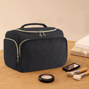 Borsa cosmetica nera trapuntata con cerniera, manico superiore, su tavolo di legno. Include pennelli trucco e palette ombretti. Accessori bellezza.