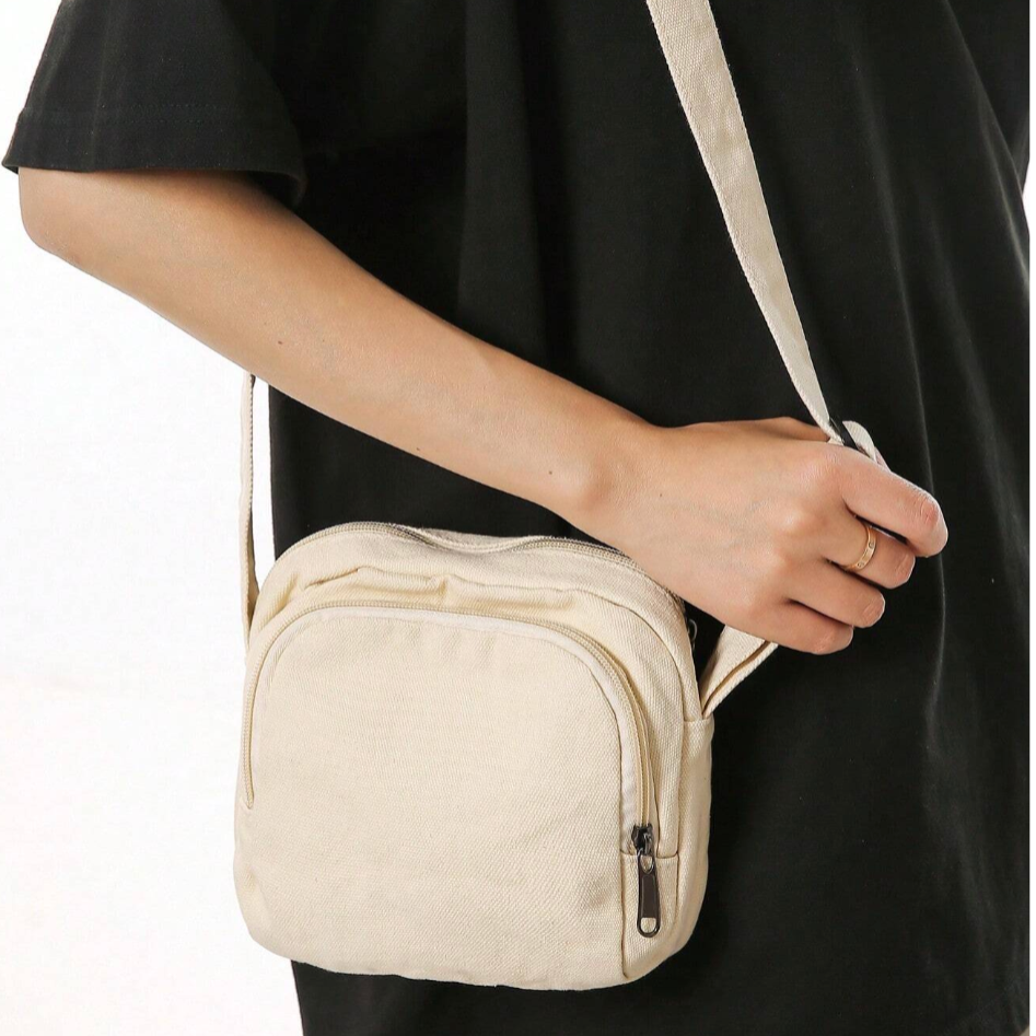 Borsa a tracolla beige in tela, indossata da una persona. Design compatto con cerniera, ideale per uso quotidiano e casual. Accessorio moda unisex.