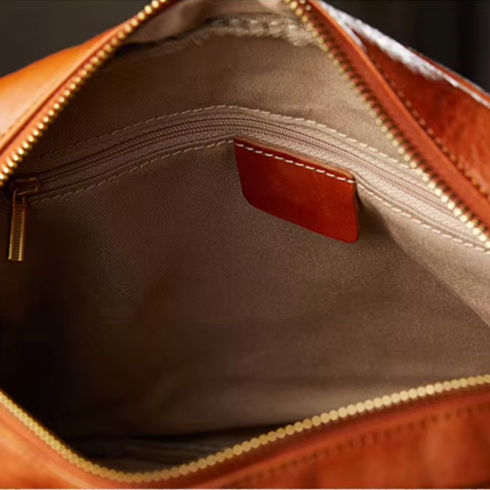 Interno di borsa in pelle marrone con cerniera aperta, tasca interna con zip e fodera beige. Accessorio moda elegante e di alta qualità.