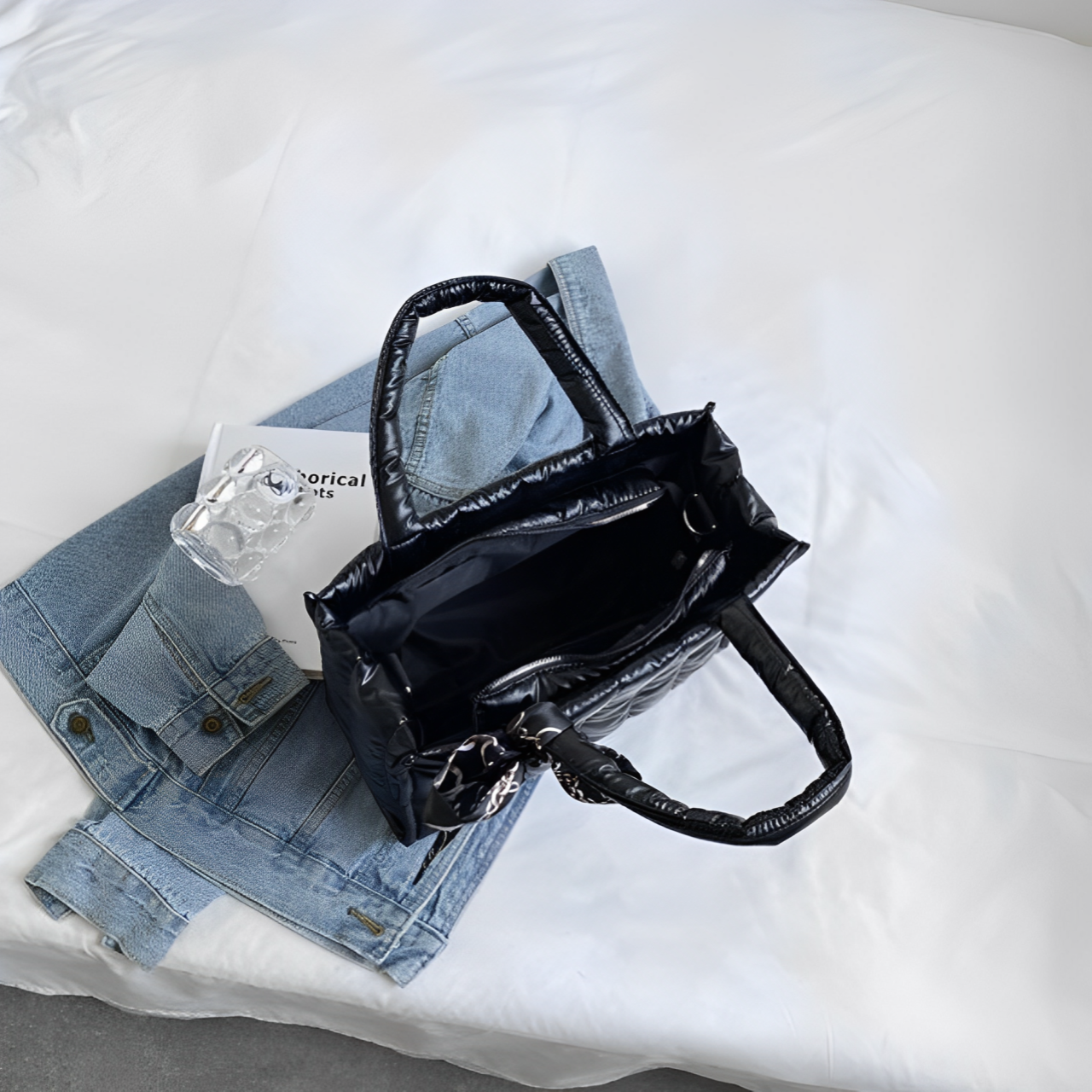 Borsa nera in pelle su letto bianco con giacca di jeans e libro. Accessori moda donna, stile casual chic.