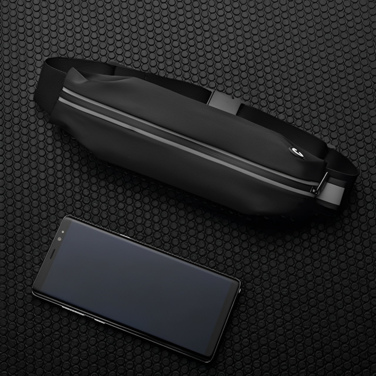 Marsupio nero impermeabile con cerniera riflettente su sfondo a nido d'ape, accanto a smartphone nero. Accessorio moda pratico e moderno.