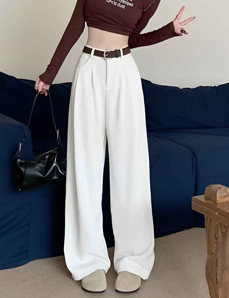 Donna in pantaloni bianchi larghi, top marrone a maniche lunghe, cintura marrone, borsa nera, posa su divano blu. Moda casual elegante.