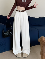 Donna in pantaloni bianchi larghi, top marrone a maniche lunghe, cintura marrone, borsa nera, posa su divano blu. Moda casual elegante.