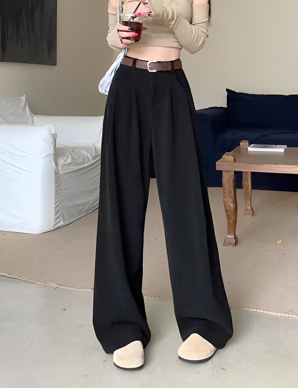 Donna con pantaloni neri a vita alta, top beige a maniche lunghe, scarpe beige, borsa bianca, in salotto moderno. Moda casual elegante.