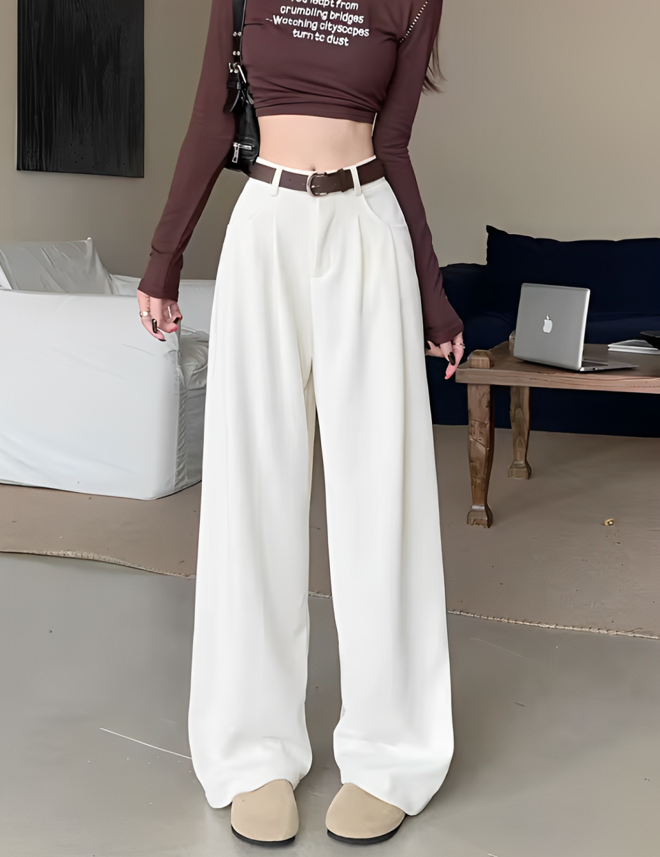Donna con pantaloni bianchi larghi, cintura marrone e top marrone a maniche lunghe. Stile moda casual, interno moderno, laptop su tavolo.