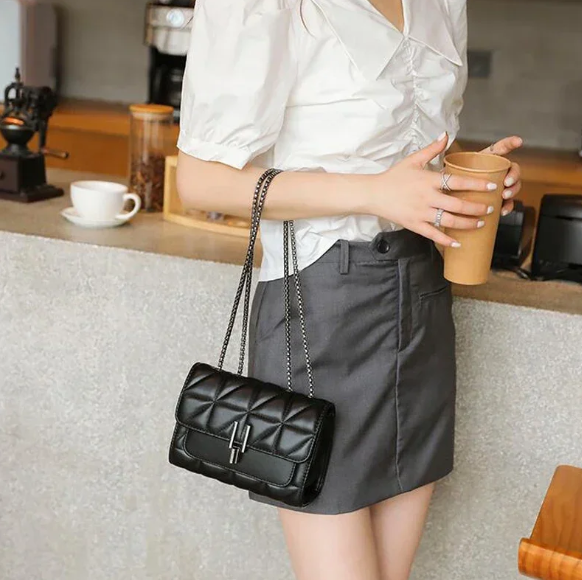 Donna con borsa a tracolla nera in pelle trapuntata, indossa camicia bianca e gonna grigia, tiene bicchiere di caffè, ambiente caffetteria.