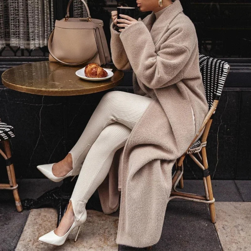 Donna elegante in cappotto beige, leggings bianchi e tacchi, seduta a un tavolo con croissant e borsa di pelle. Moda autunnale chic.