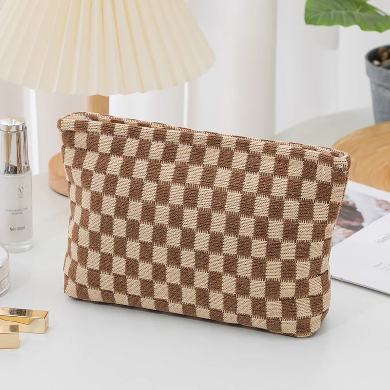 Borsa a scacchi marrone e beige su tavolo bianco, accanto a lampada e piante. Design elegante, ideale per moda e accessori.