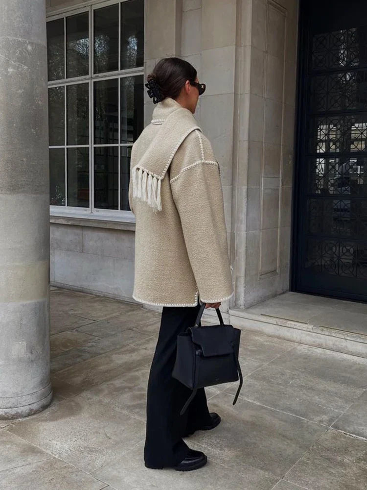 Donna con cappotto beige oversize, pantaloni neri e borsa a mano nera, cammina su marciapiede urbano. Stile autunnale elegante e moderno.