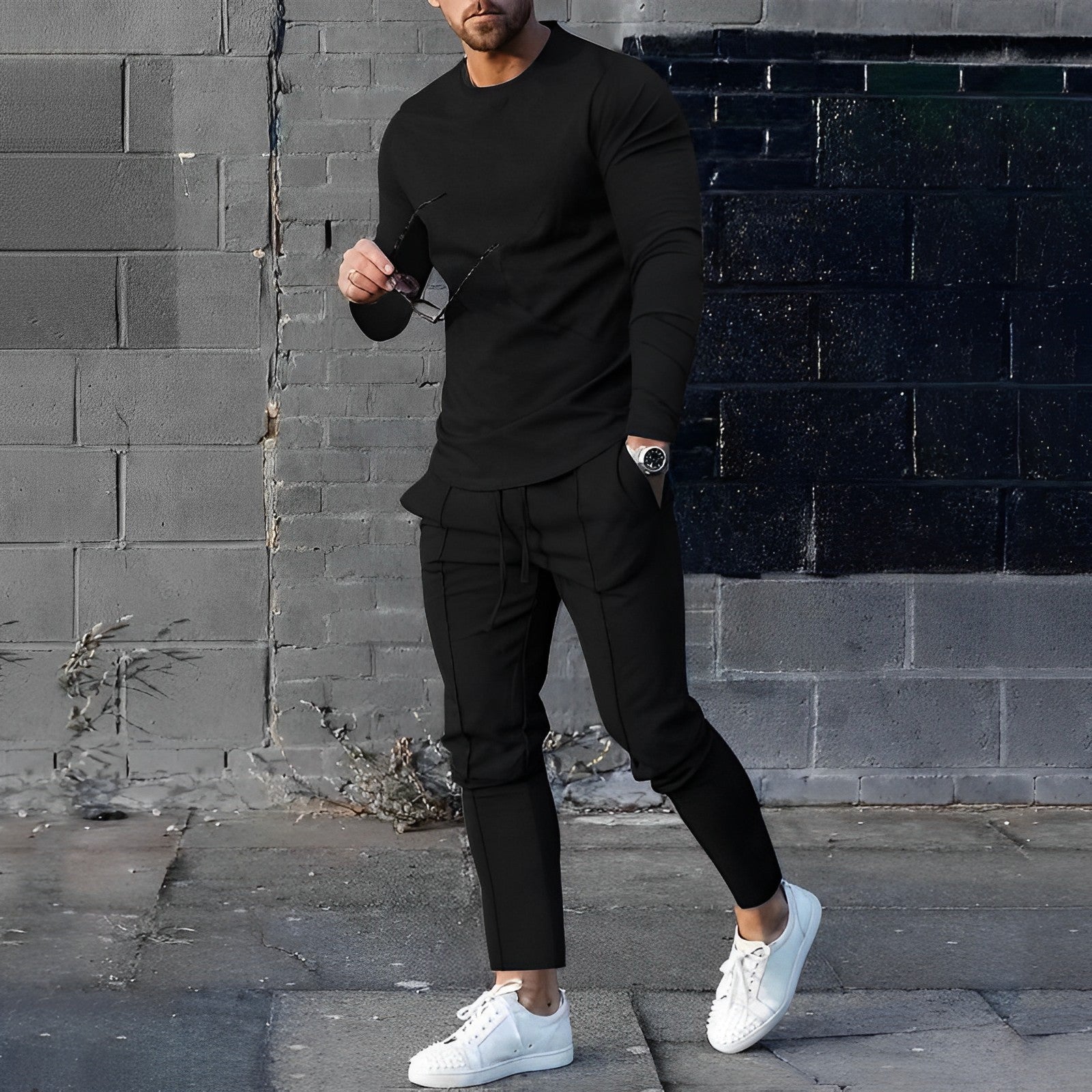 Uomo in abbigliamento casual nero, maglia e pantaloni, con scarpe bianche, posa davanti a un muro di mattoni. Moda maschile urbana elegante.