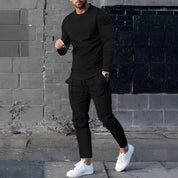 Uomo in abbigliamento casual nero, maglia e pantaloni, con scarpe bianche, posa davanti a un muro di mattoni. Moda maschile urbana elegante.