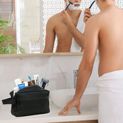 Uomo che si rade davanti allo specchio con rasoio, accanto a beauty case nero con prodotti da bagno. Bagno moderno, cura personale maschile.