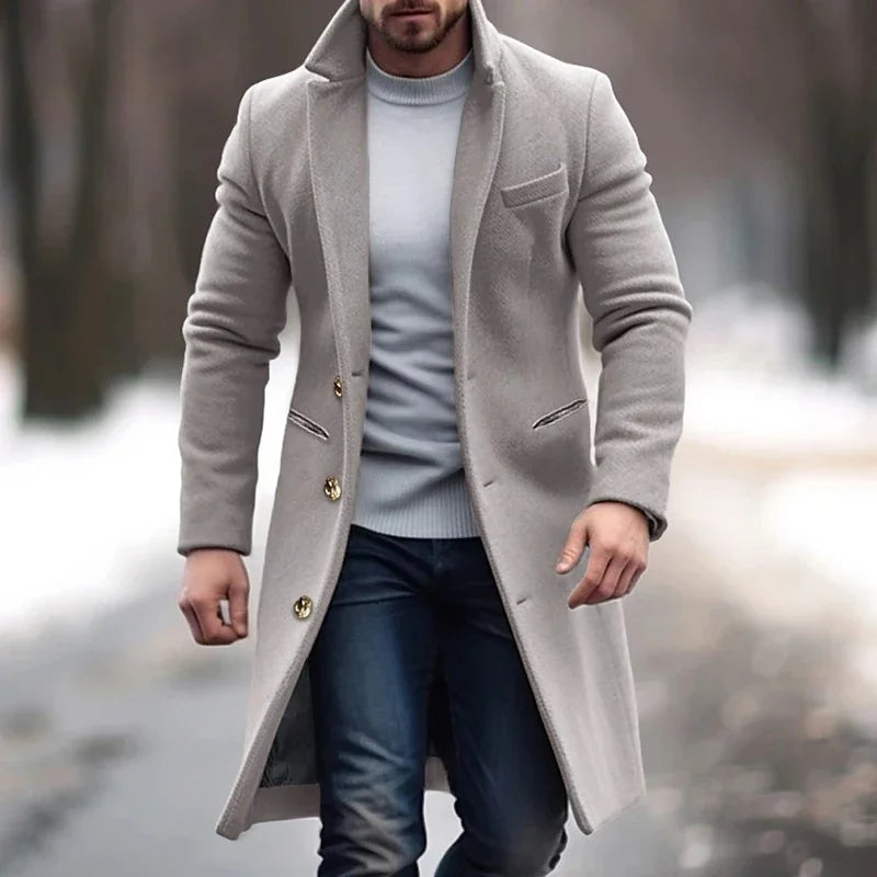 Cappotto uomo elegante grigio chiaro, lungo, con bottoni dorati, indossato su maglione bianco e jeans. Moda invernale maschile.