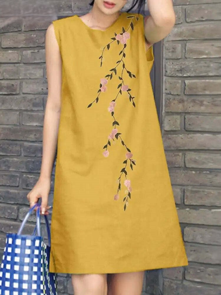 Abito estivo giallo senape senza maniche con motivo floreale ricamato, donna con borsa a quadri blu, sfondo muro di mattoni.