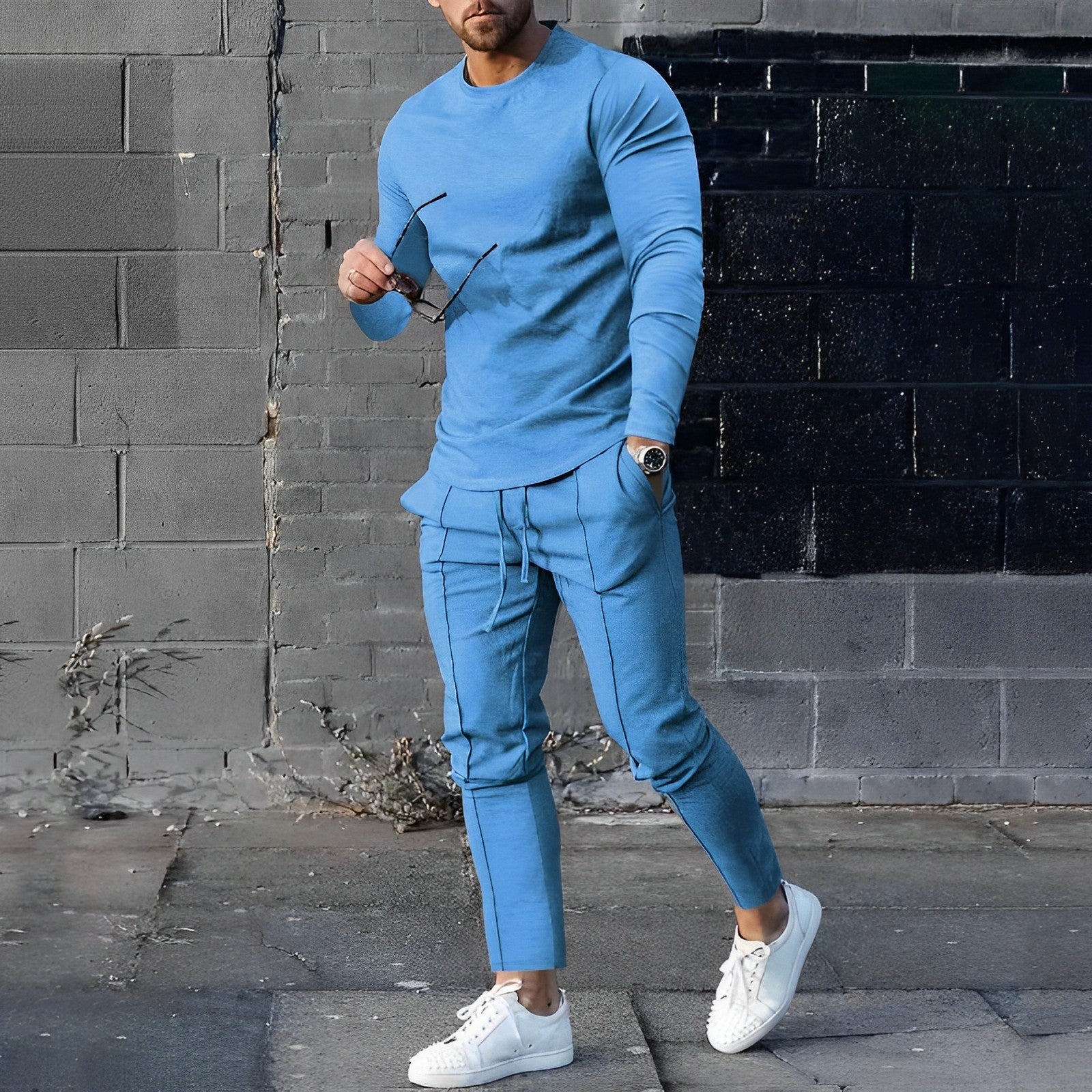 Uomo in tuta sportiva blu, con occhiali da sole in mano, cammina su marciapiede. Indossa scarpe bianche. Look casual e moderno.