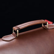 Manico di valigetta in pelle marrone con cuciture bianche, dettagli in metallo dorato, sfondo nero. Elegante accessorio business.
