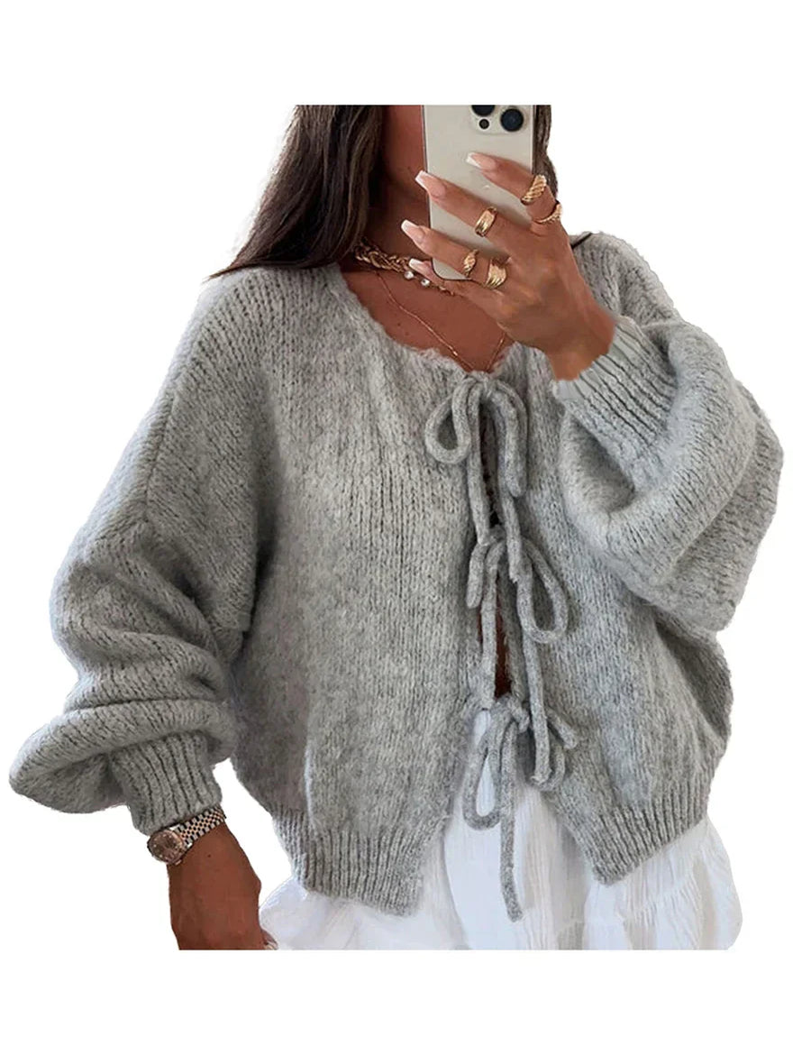 Donna con cardigan grigio in maglia morbida, maniche lunghe, dettagli a fiocco, abbinato a gonna bianca. Stile casual chic, moda autunnale.