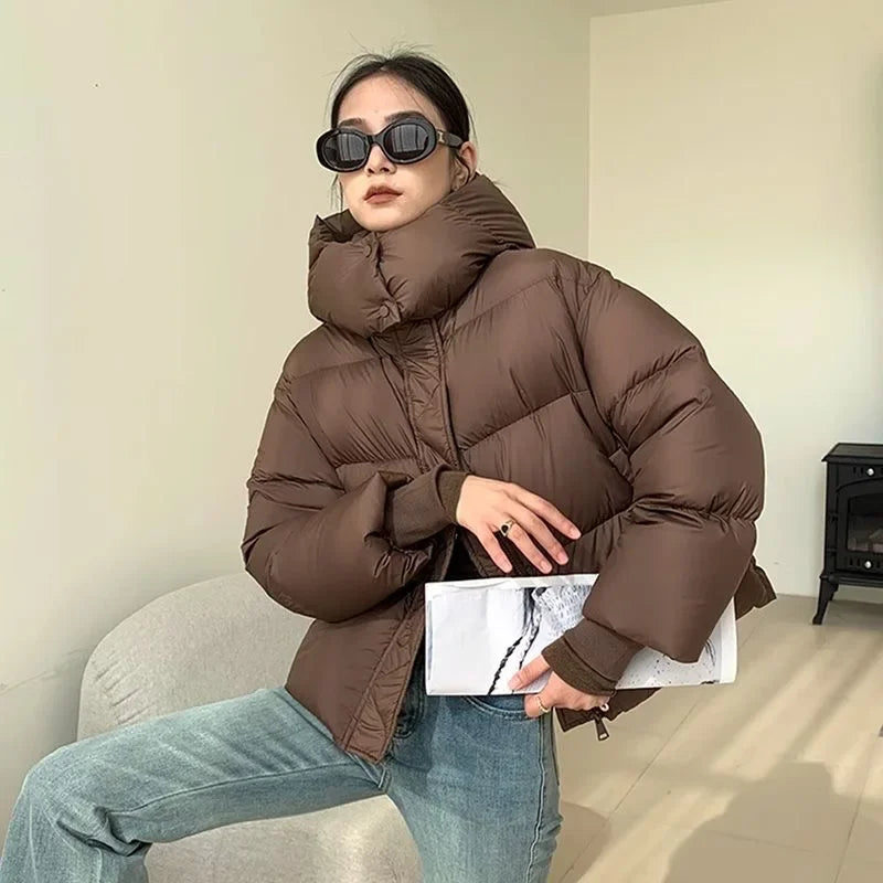 Giacca invernale donna marrone, imbottita, con collo alto. Moda autunno-inverno, stile casual. Occhiali da sole neri, jeans chiari.