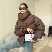 Giacca invernale donna marrone, imbottita, con collo alto. Moda autunno-inverno, stile casual. Occhiali da sole neri, jeans chiari.
