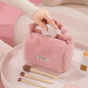 Borsa cosmetica rosa in pelliccia sintetica con manico, circondata da pennelli trucco su sfondo rosa. Accessorio moda elegante e pratico.