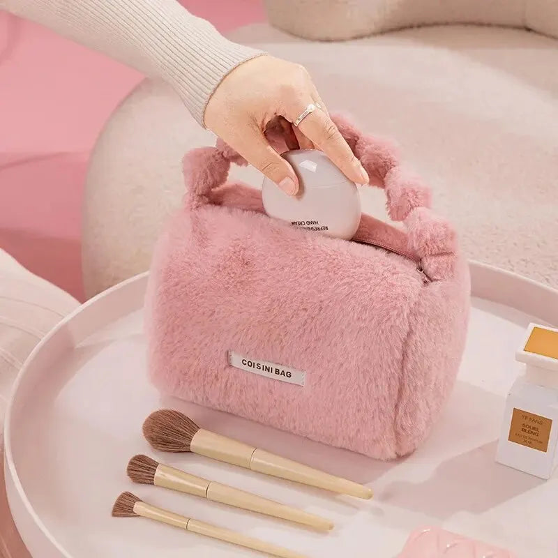 Borsa cosmetica rosa in pelliccia sintetica con manico, circondata da pennelli trucco su sfondo rosa. Accessorio moda elegante e pratico.