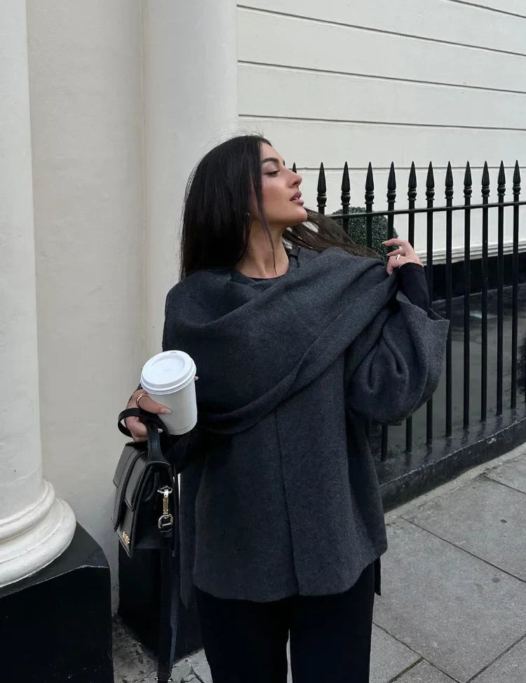 Donna con maglione grigio oversize, sciarpa abbinata, borsa nera e caffè da asporto. Moda autunnale elegante, look urbano casual.
