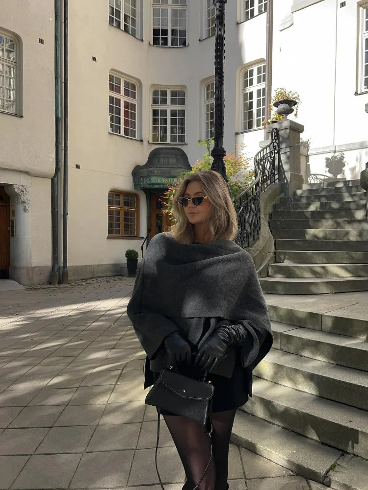 Donna elegante con occhiali da sole, mantella grigia e guanti neri, posa su una scalinata storica in città europea. Moda autunnale urbana.