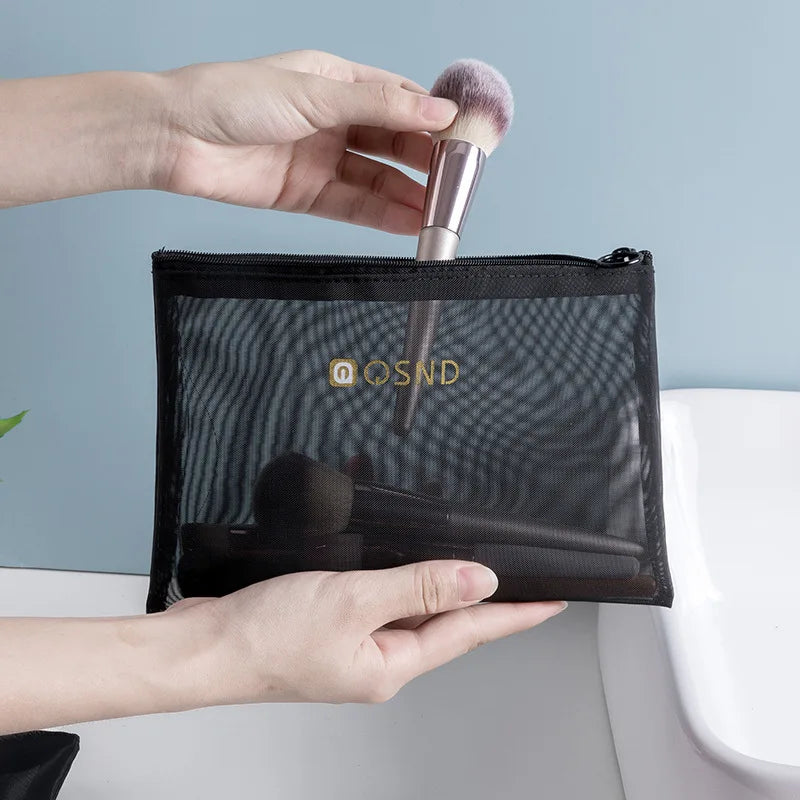 Mani che inseriscono un pennello da trucco in una pochette trasparente nera con cerniera, ideale per organizzare cosmetici e accessori da viaggio.